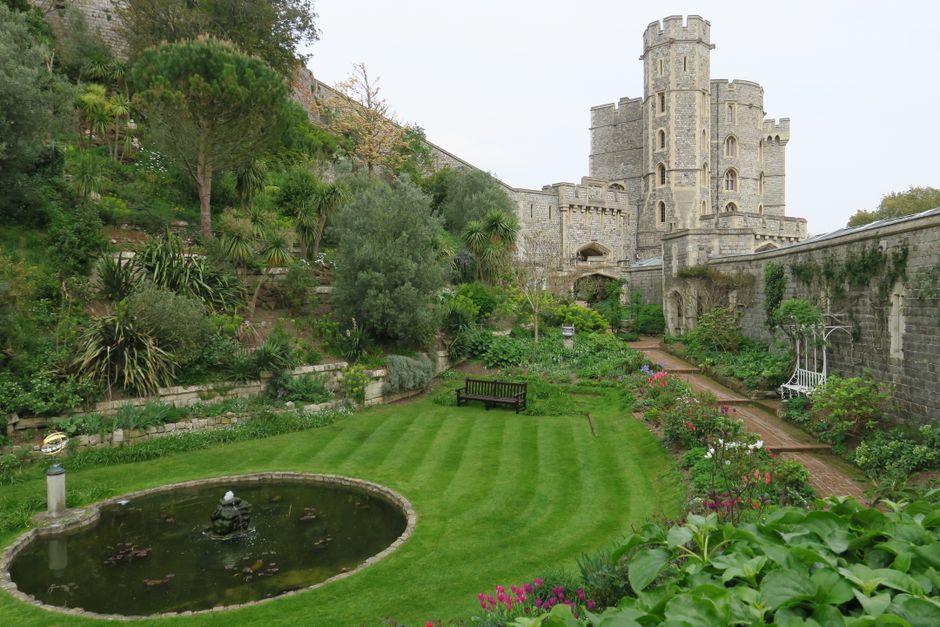 A picturesque garden with neatly trimmed, vibrant green lawn featuring fresh, striped grass patterns, bordered by flower beds with blooming roses and various lush plants. A small circular pond with wa
