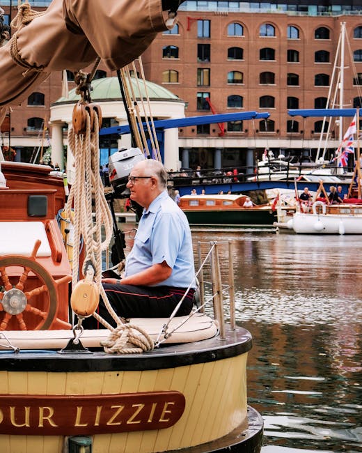 A man with gray hair and glasses, dressed in a light blue short-sleeved shirt, is seated on the deck of a small wooden boat named 'Our Lizzie' docked in a marina. The boat features a cream and black h